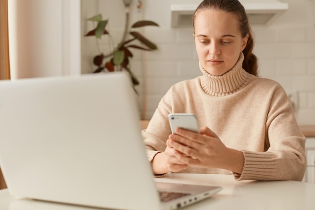 Horizontal shot of young adult beautiful woman wearing beige jumper posing in kitchen and working on laptop sitting at table and typing on smart phone