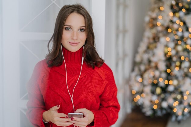 Horizontal shot of young Caucasian woman enjoys music from playlist listens favourite song with earphones dressed in knitted red sweater stands against decorated Christmas tree background