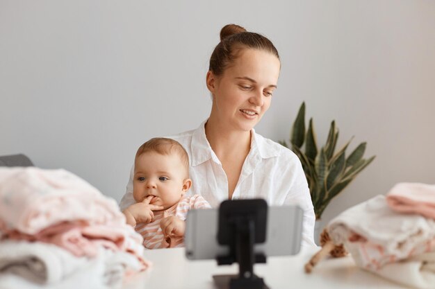 Horizontal shot of young female with hair bun broadcasting livestream at home, together with her infant daughter, filming content for her blog, talking about mom's problems in raising children.