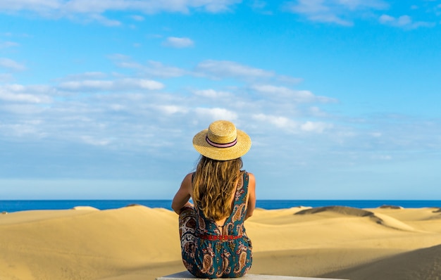 Ripresa orizzontale di una giovane donna con un cappello di paglia in piedi su una spiaggia sabbiosa sotto un cielo limpido e luminoso