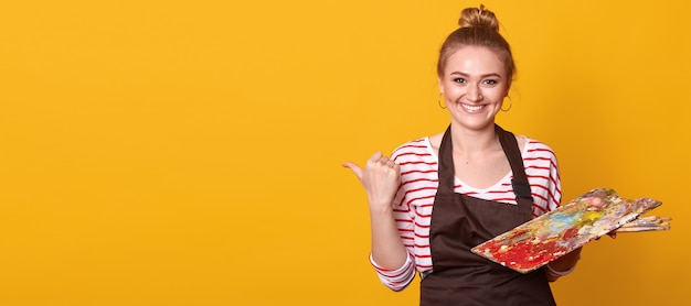 Horizontal shot of young smiling blonde woman artist isolated over yellow studio, holding brushes and palette of colors. Paintings and art equipment concept.