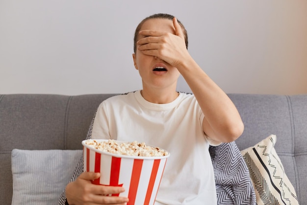 Horizontal shot of young woman wearing white t shirt sitting on\
sofa with popcorn and remote control watching thriller with scared\
episode covering eyes with hand