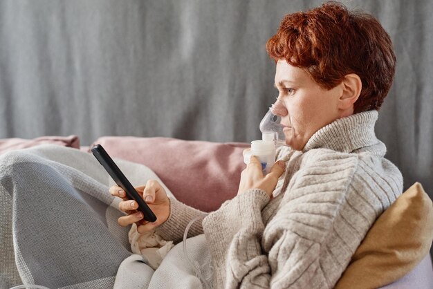 Horizontal side view shot of mature woman suffering cold staying at home relaxing on sofa using nebu