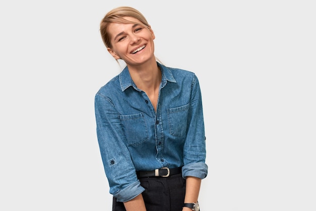 Horizontal studio portrait of happy blonde European woman smiling broadly standing in blue denim shirt over white background Business female posing on studio wall with copy space for your promotion