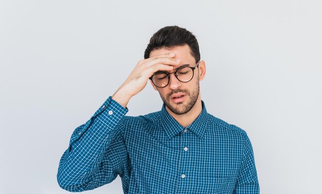 Horizontal studio portrait of male in blue shirt has headache with hand on forehead looking down after overworking isolated on white wall background with copy space People health emotion concept