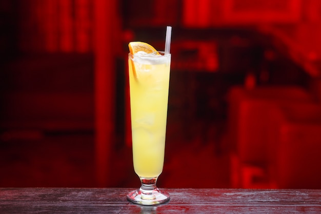 Horizontal view. Closeup of a long glass of orange juice with gin, which sits on the bar counter, isolated on a red light space.