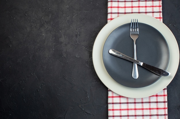 Horizontal view of cutlery set on dark gray color and white empty plates on red stripped towel on the left side on black background with free space