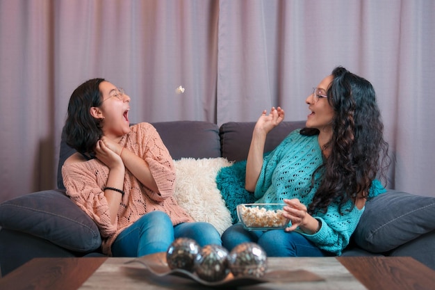 Horizontal view of a mom and her daughter watching a movie at home they play throwing popcorn to catch it with their mouths they make very funny expressions