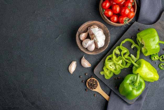 Horizontal view of whole cut chopped green peppers on wooden cutting board tomatoes in bowl garlics on dark color towel on the left side on black surface