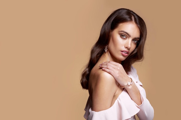 Horizontal view of a young woman with bare shoulder, brown wavy hair, model charm with bracelet and earrings over brown background. Beauty people portrait. Copy space.