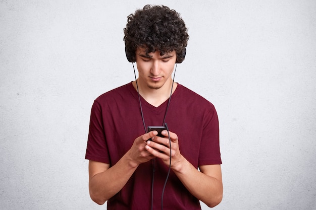 Horizontal young man messaging and blogging on cellphone while listening music in earphone. Handsome male posing on wall with blank copy space. People and technology concept.