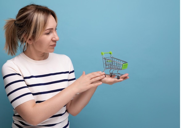 Horizontale foto van een jonge vrouw die shopaholic is met een winkelwagentje op een blauwe achtergrond met kopie