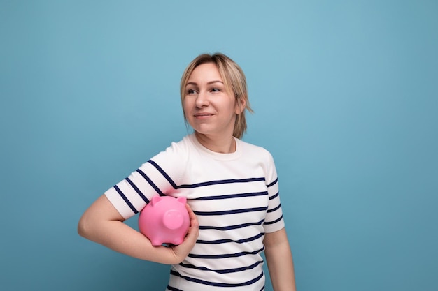 Horizontale foto van een peinzende aantrekkelijke jonge vrouw in een casual outfit met een roze spaarvarken van