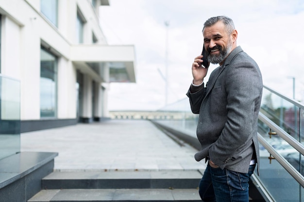 Horizontale foto van een volwassen volwassen mannelijke baas met grijs haar en baard die aan de telefoon praat op de