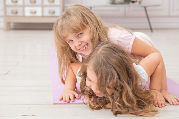Horizontale foto van paar kleine meisjes spelen op yoga mat glimlachend in de woonkamer, gezonde levensgewoonten en levensstijl concept.