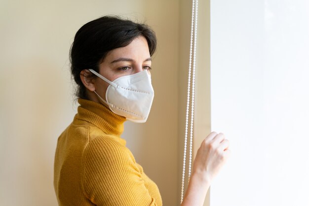 Horizontale mening van zieke vrouw die een gezichtsmasker draagt dat door het venster kijkt.