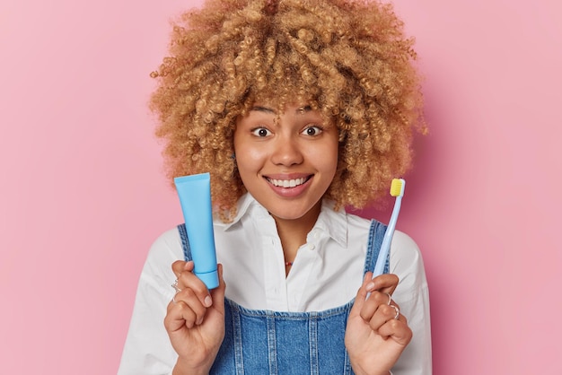 Horizontale opname van vrolijke vrouw zorgt voor mondhygiëne borstels tanden houdt regelmatig een tube tandpasta vast en tandenborstel draagt een wit overhemd en denim sarafan geïsoleerd over roze achtergrond