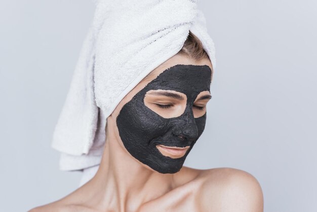 Horizontale portret van mooie jonge vrouw met klei gezicht zwart masker doen wellness therapie thuis poseren tegen witte muurKaukasische vrouw organische gezichtsmasker met witte handdoek op hoofd toe te passen