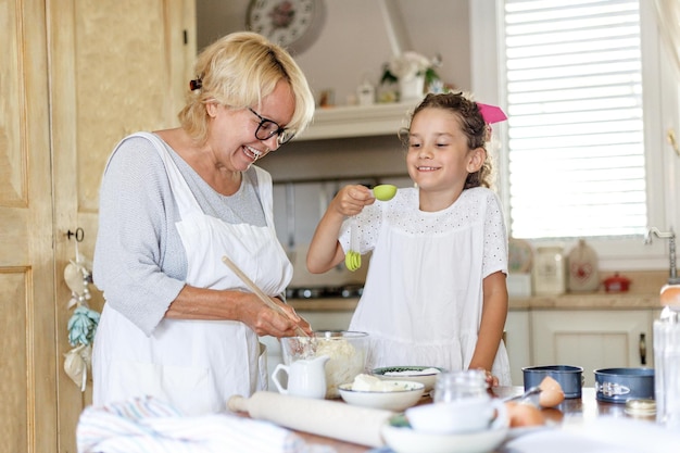 Horizontale weergave. Oma en nichtje koken in de keuken, kneed een deeg in een kom, nichtje voegt suiker toe met een theelepeltje.