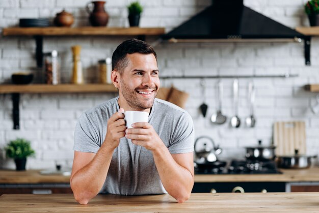 Horizontale weergave van de knappe man zittend op de stoel leunde op tafel, glimlachend en wegkijkend terwijl hij koffie dronk. Beeld bij de gezellige keuken