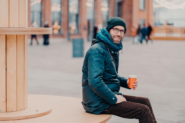 Horizontale weergave van vrolijke Europese man met dikke haren draagt hoed en jas houdt wegwerp kopje koffie ademt frisse lucht poses op straat Mannelijk model geniet van verse drank buiten