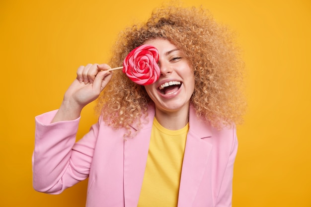 Horizotal shot of curly happy woman covers eye with tasty sweet candy has fun laughs happily dressed in formal clothes isolated over vivid yellow wall. Lovely glad female holds lollipop