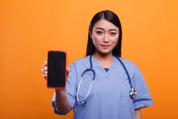 Hospital caregiver wearing medical instrument while holding mobile cellphone on orange background. Attractive healthcare clinic nurse wearing stethoscope while holding modern smartphone device.