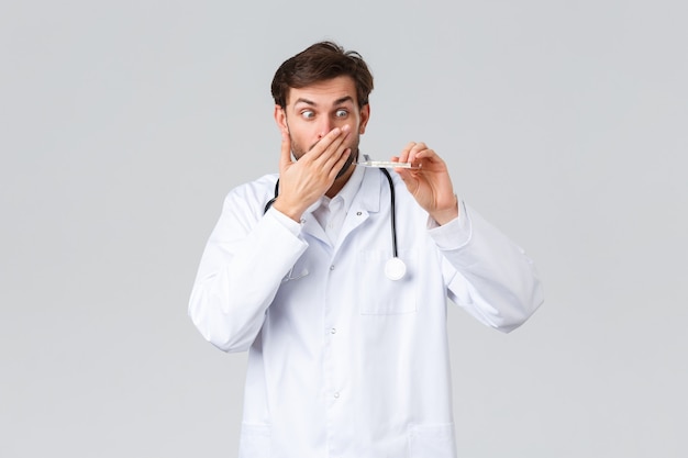 Hospital, healthcare workers, covid-19 treatment concept. Shocked and concerned doctor in white scrubs with stethoscope, gasping, stare at thermometer showing high fever, grey background