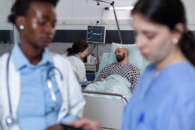 Hospital resident consulting patient connected to monitor measuring vitals and pulse oximeter while doctor and nurse look at digital tablet. Team of doctors consulting man with lung problems.