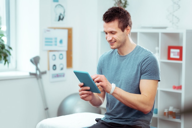 In the hospital room. Positive young man using his tablet while waiting for a doctor in the hospital room