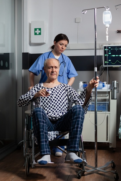 Hospitalized senior man sitting in wheelchair in hospital room, holding iv drip with oximeter attached on finger