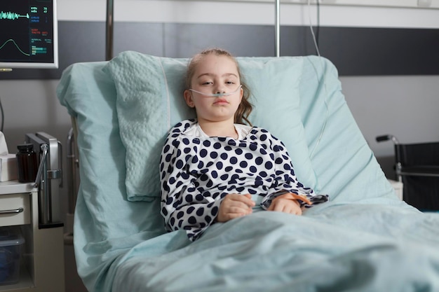 Госпитализированная больная маленькая девочка отдыхает в детской поликлинике. Под лечением больной ребенок лежит в педиатрической палате больницы и смотрит в камеру.