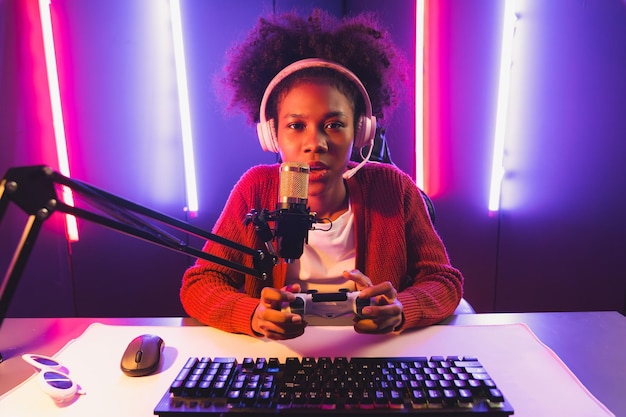 Photo host channel of gaming streamer african girl playing online game with joystick talking with viewers media online on microphone esport skilled team players in neon color lighting room tastemaker