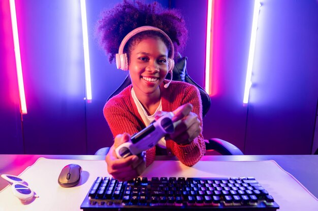 ゲームストリーマーのホストチャンネル ジョイスティックでオンラインゲームをプレイするアフリカの女の子がマイクでオンラインで視聴者メディアと話す ネオンカラー照明室でEスポーツの熟練したチームプレーヤー