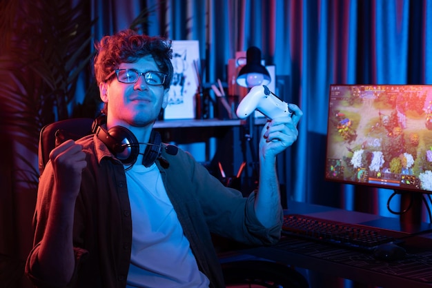 Host channel of young gaming streamer looking at camera holding joystick with fighting Moba battle arena game with multiplays team against on pc screen background at neon digital light room Gusher