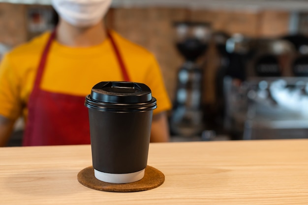 Tazza di caffè nero caldo sul bancone con il personale della cameriera che indossa una maschera protettiva sullo sfondo in caffetteria, consegna cibo, ristorante caffetteria, cibo da asporto, piccolo imprenditore, concetto di cibo e bevande