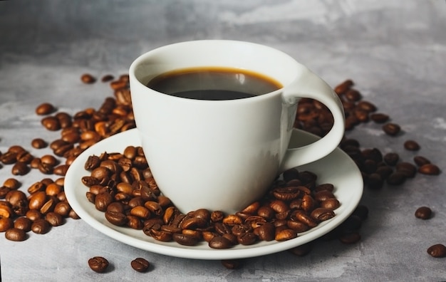 Hot black coffee in white ceramic cup with coffee beans around on textured grey background