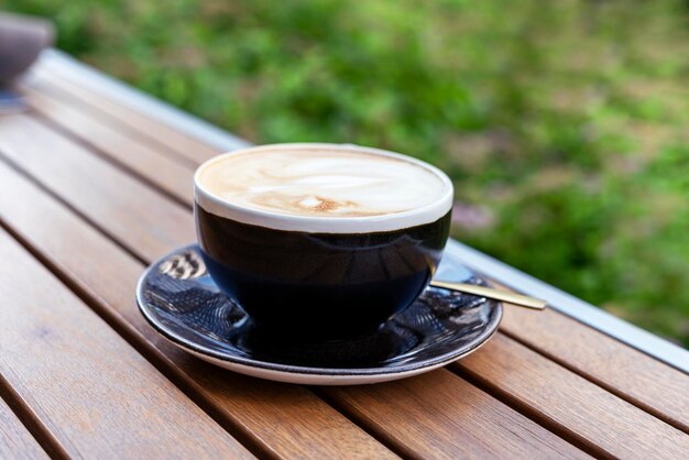 hot cappuccino coffee in black cup on wooden table on summer terrace against backdrop of green plant