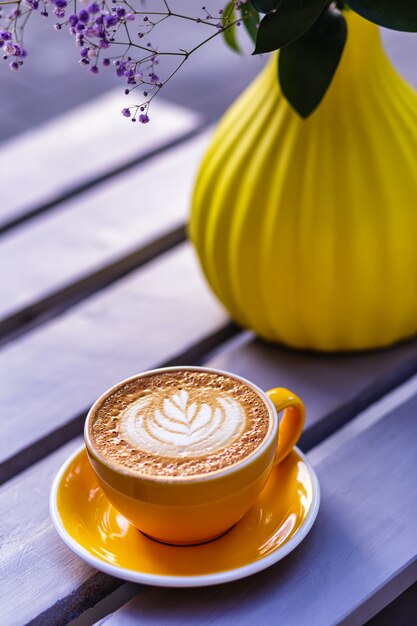 Hot cappuccino coffee in an orange cup a yellow vase with flowers in the background