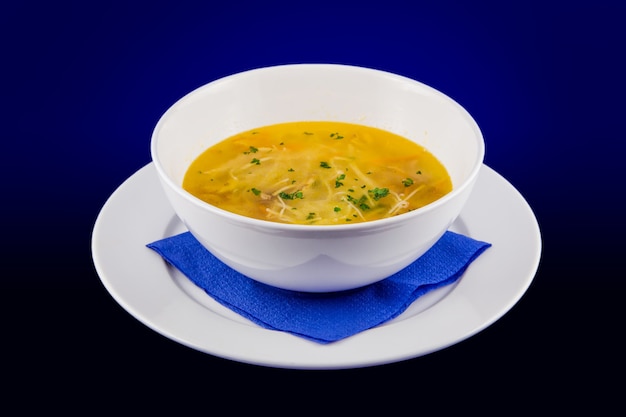 Горячий куриный суп с лапшой с морковью и зеленью. Крупный план на белой тарелке на синем фоне.