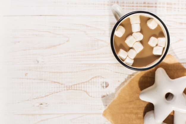 Hot Christmas drink with marshmallows in an iron mug and gingerbread cookies, on a white table.