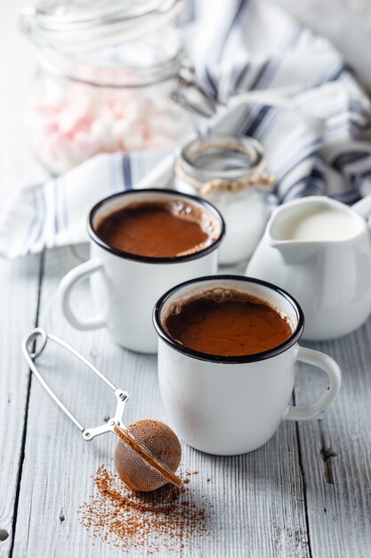 Hot cocoa in two enameled white mugs on a light wooden table.