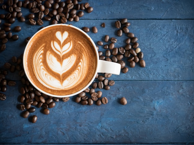 Hot coffee in a cup with foam milk beautiful latte art and coffee beans on blue wooden table background. Cappuccino or latte with froth foam, drinks concept.