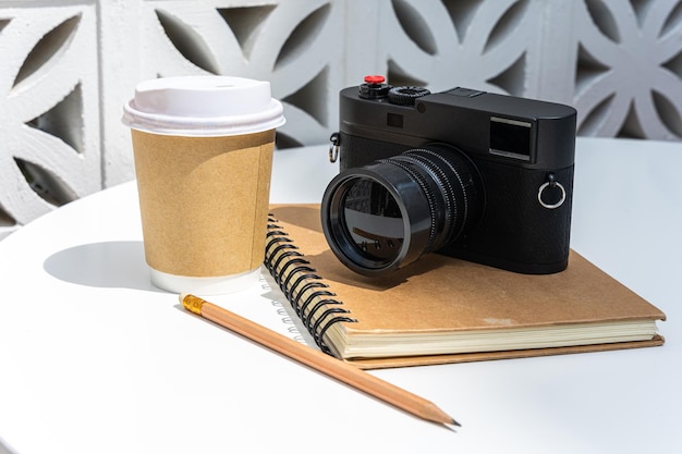 紙で作られたカップにラテを入れたホットコーヒーラテと、ビジネスワークコンセプト中のカフェの喫茶店での朝食として、白い木のテーブルの上にノートにカメラ