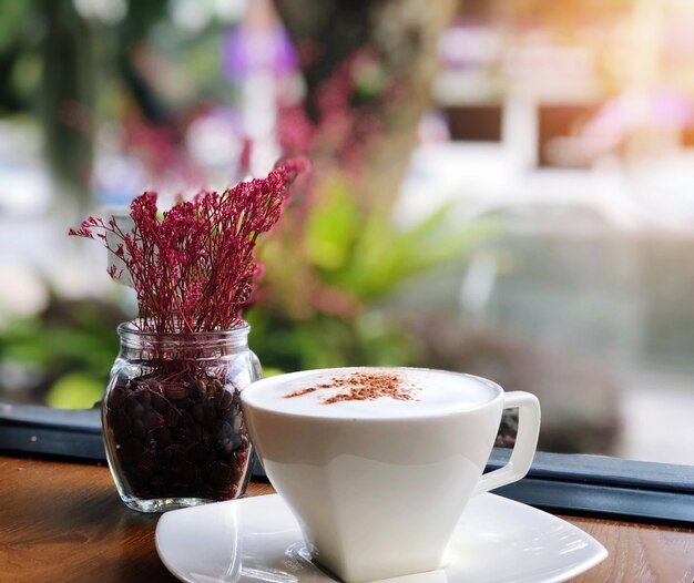 Hot Coffee in a white cup on wooden table and flowers vase in Coffee shop blur background