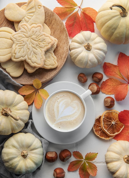 Hot coffee with cookies in a white cup surrounded by autumn leaves and pumpkins on a white wooden background
