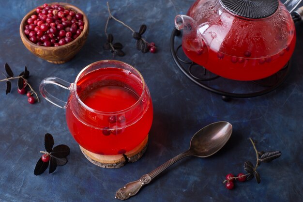 Tè al mirtillo rosso caldo in una tazza di vetro e una teiera su uno sfondo scuro e freddo.