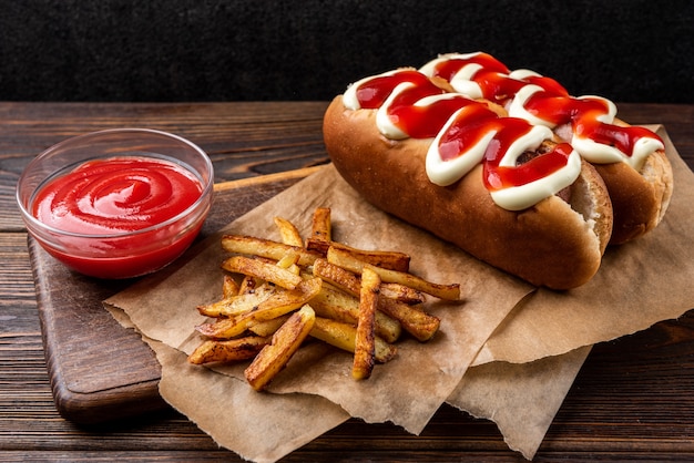 Hot dog with mayonnaise and ketchup and French fries on dark wooden background.