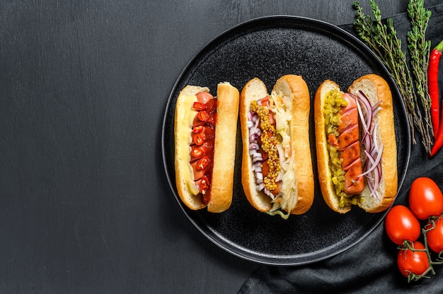 Hot dogs fully loaded with assorted toppings on a tray. Delicious hot-dogs with pork and beef sausages. Black background. Top view. Copy space