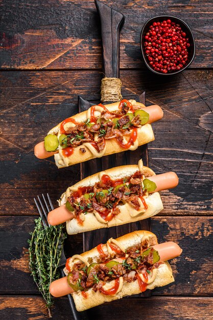 Hot dogs fully loaded with fried bacon, onion and pickled cucumbers. Dark wooden background. Top view.